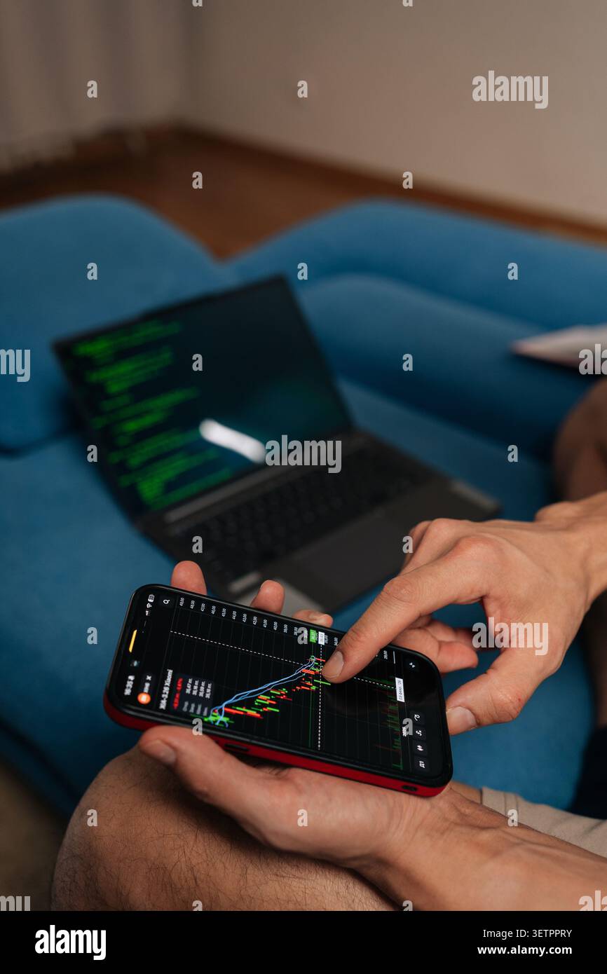 Hands holding smartphone with financial charts while laptop showing code rests on blue sofa, illustrating remote work, trading, coding, data-driven Stock Photo