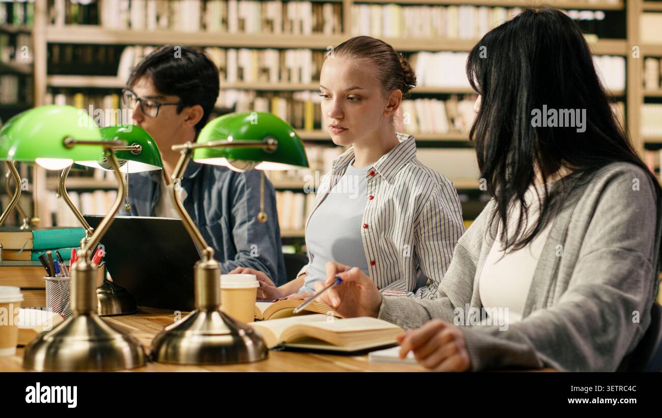 University students discussing with tutor and studying with open books at library desk, taking notes and preparing for coursework. Study group using academic study materials, higher education scene. Stock Photo