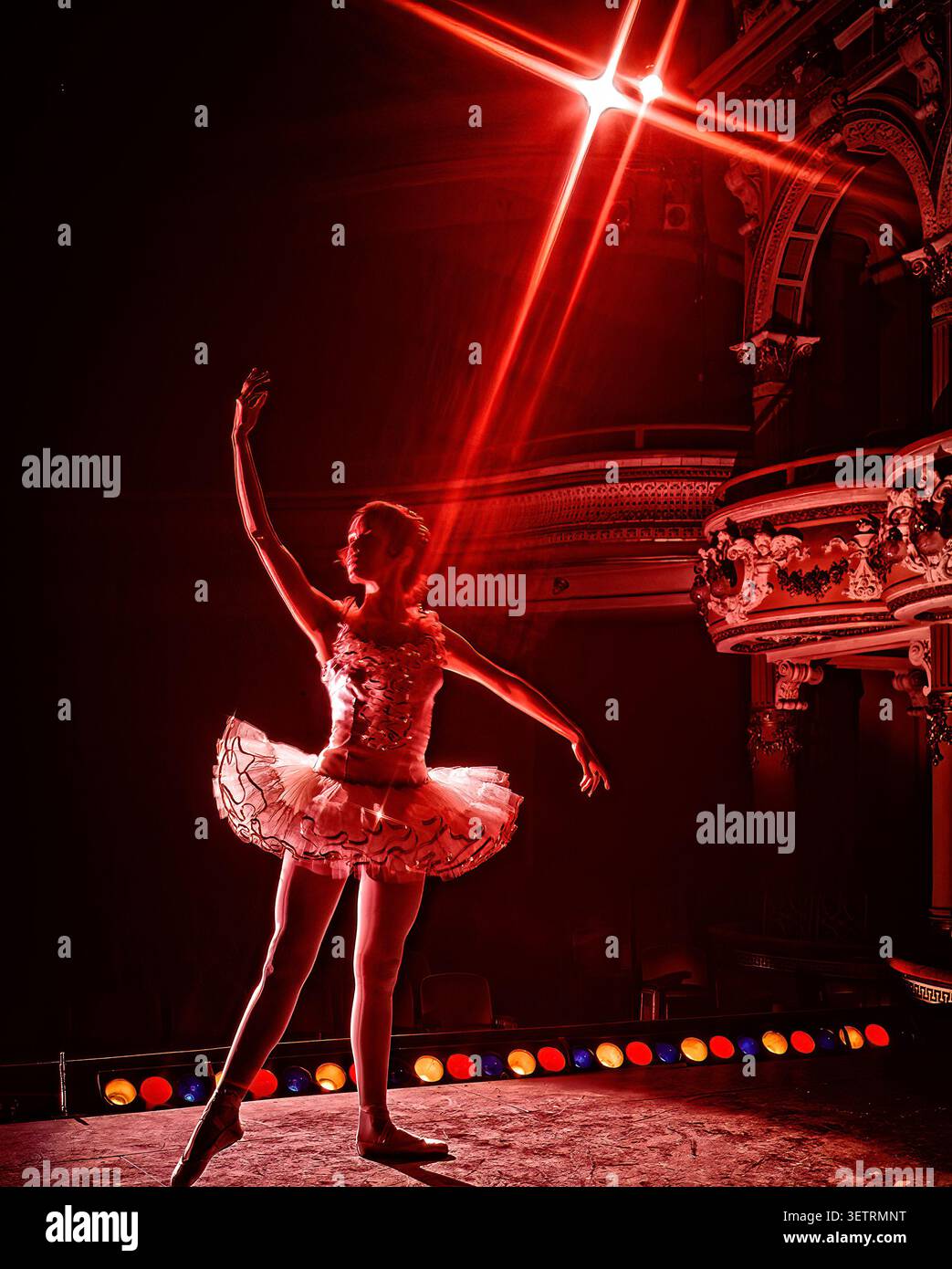 Ballerina poised beneath  radiant red stage light inside ornate theater, dramatic lens flare, architectural elegance creating striking performance Stock Photo