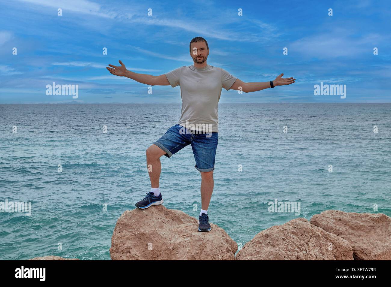 A young man stands on a stone against the background of the sea with his arms outstretched Stock Photo