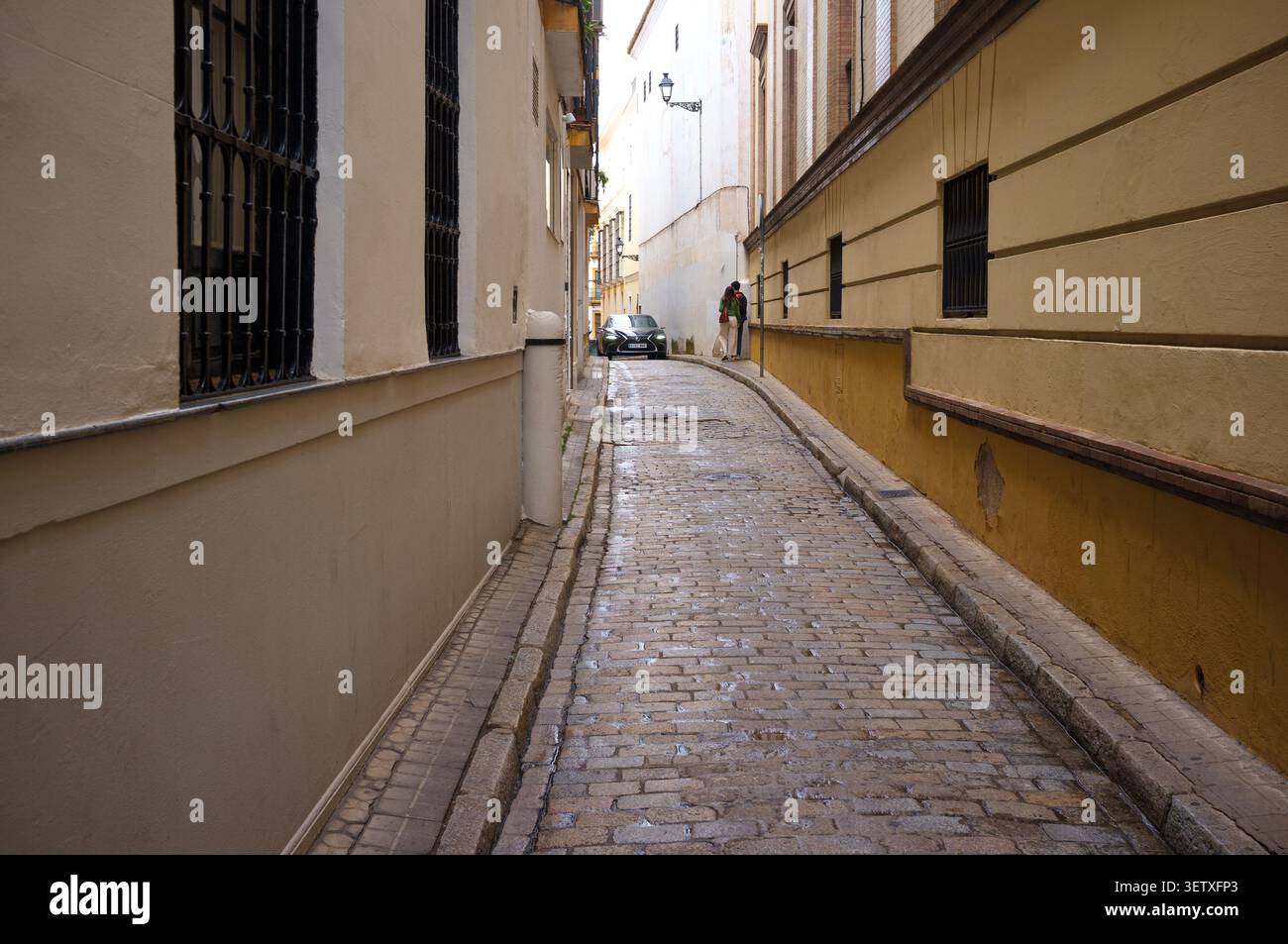 The tight narrow cobbled streets of Seville Spain with a car driving down the road in shadow showing the natural cooling effect of close buildings Stock Photo