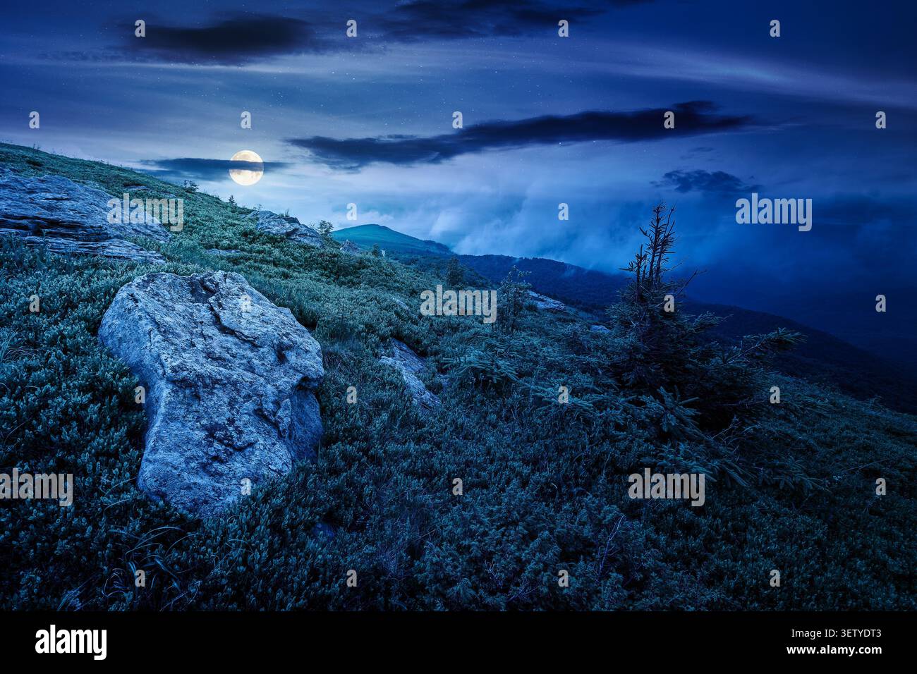 green landscape of mountain in summer at night. mysterious view of rock among grass on the hillside. full moon over travel background under a dark sky Stock Photo