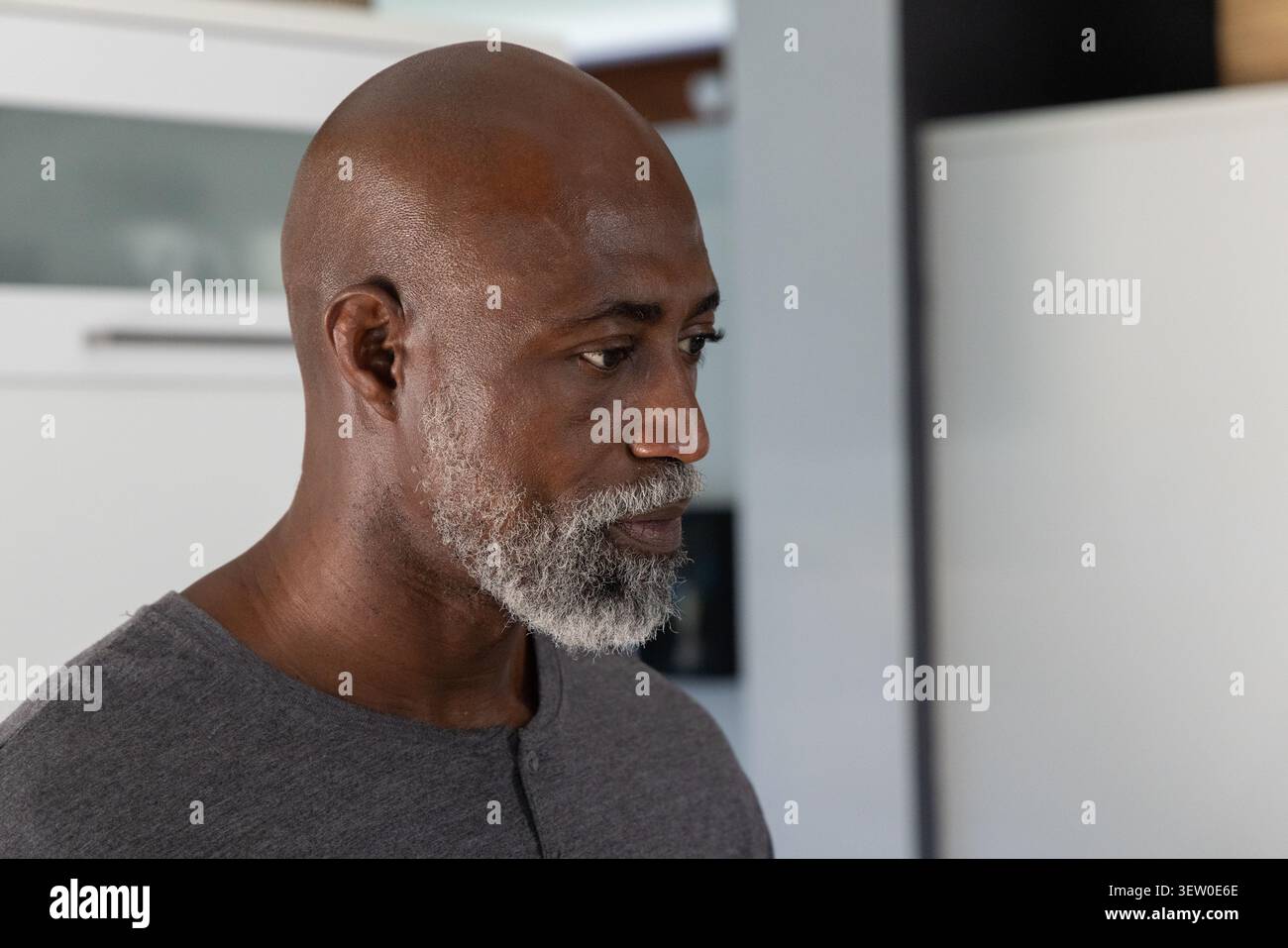 African American man standing, looking right in kitchen with white cabinets, wearing gray shirt Stock Photo