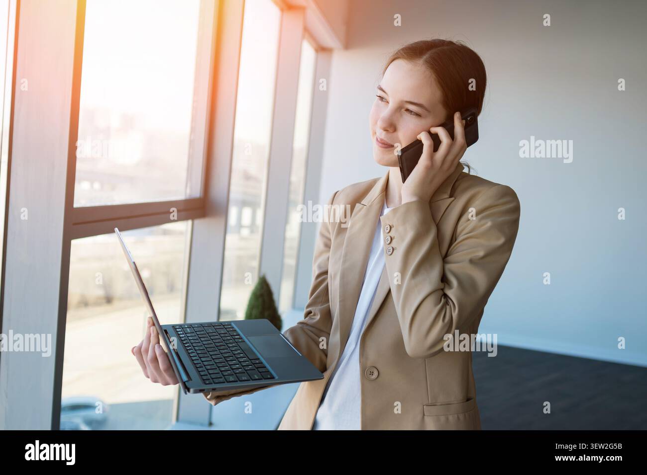 Professional businesswoman multitasking with laptop and smartphone by window Stock Photo