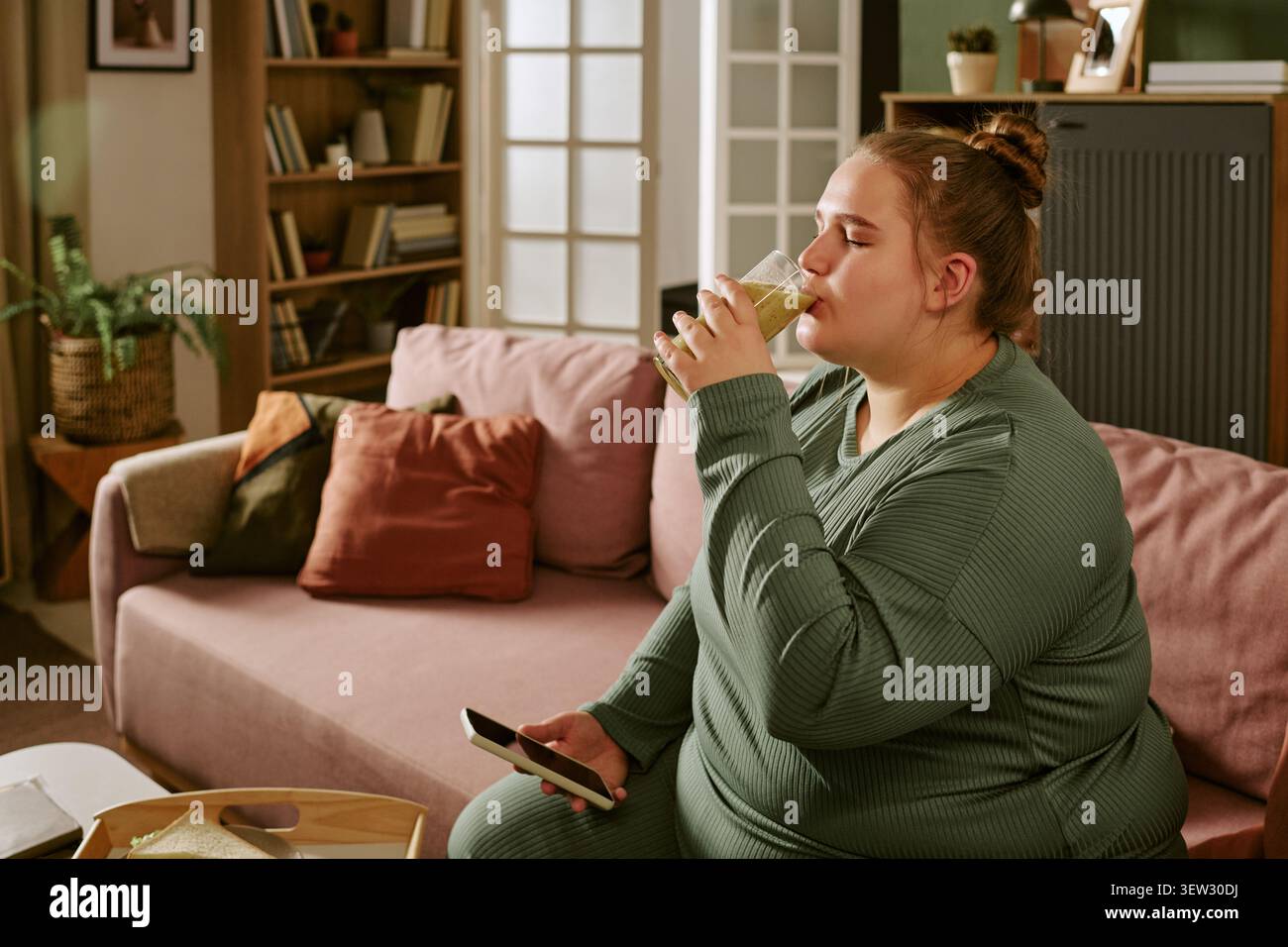 Plus Size Woman Drinking Smoothie While Using Smartphone at Home Stock Photo