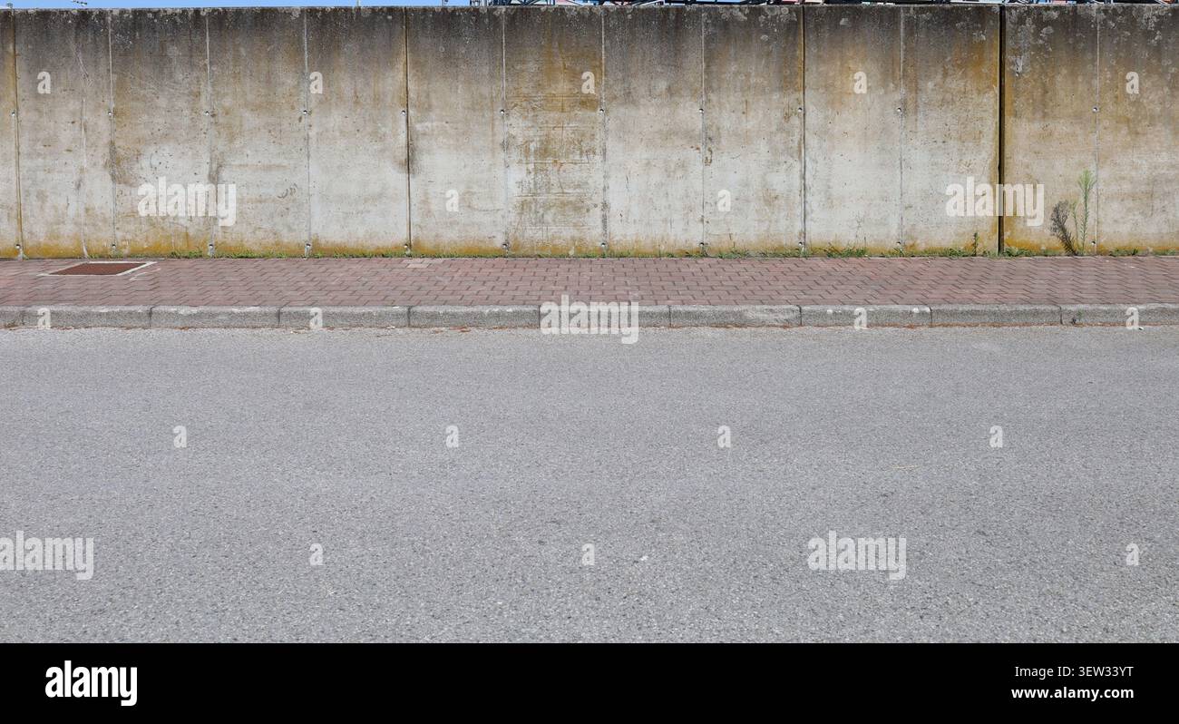 Grunge prefab concrete boundary wall with with weathering marks, stains, moss, and lichens. Sidewalk and street in front. Background for copy space Stock Photo