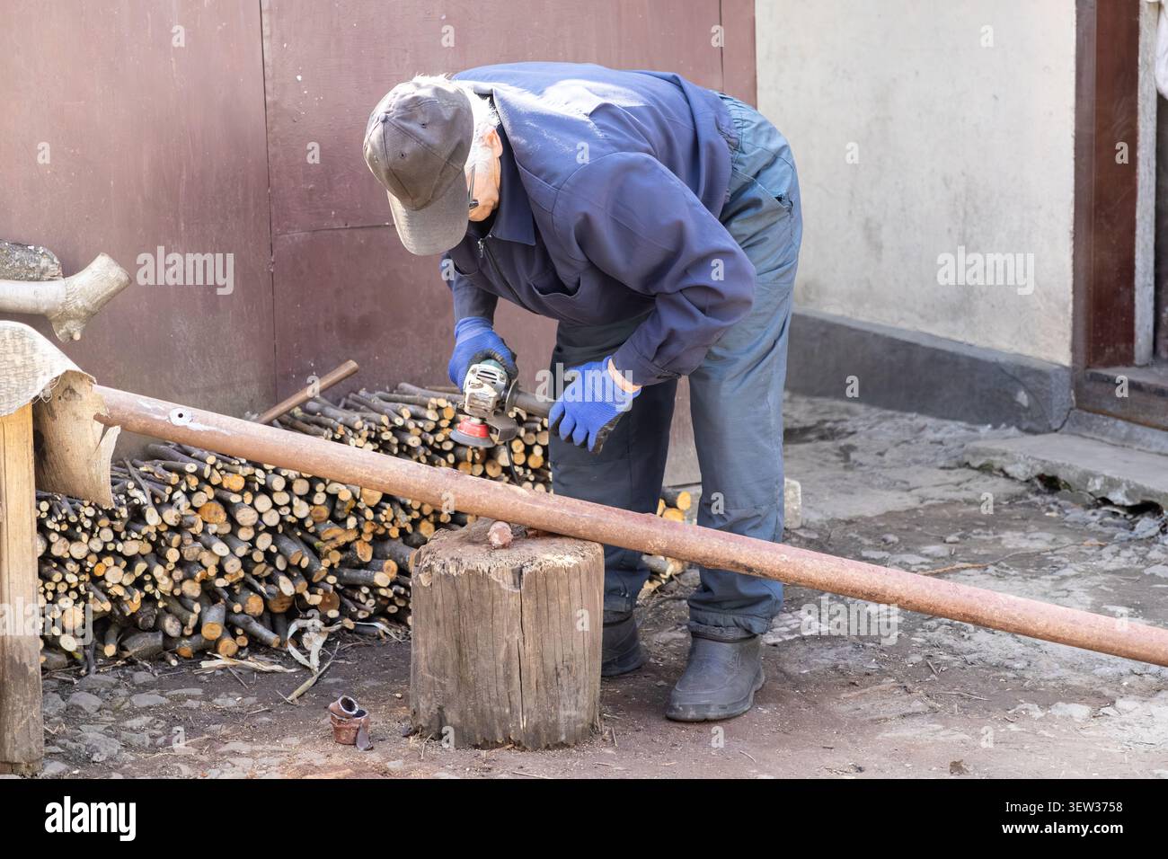 A skilled craftsman cuts a metal pipe with a power tool on a wooden block at a rural home site, demonstrating professional repair work, technical skil Stock Photo