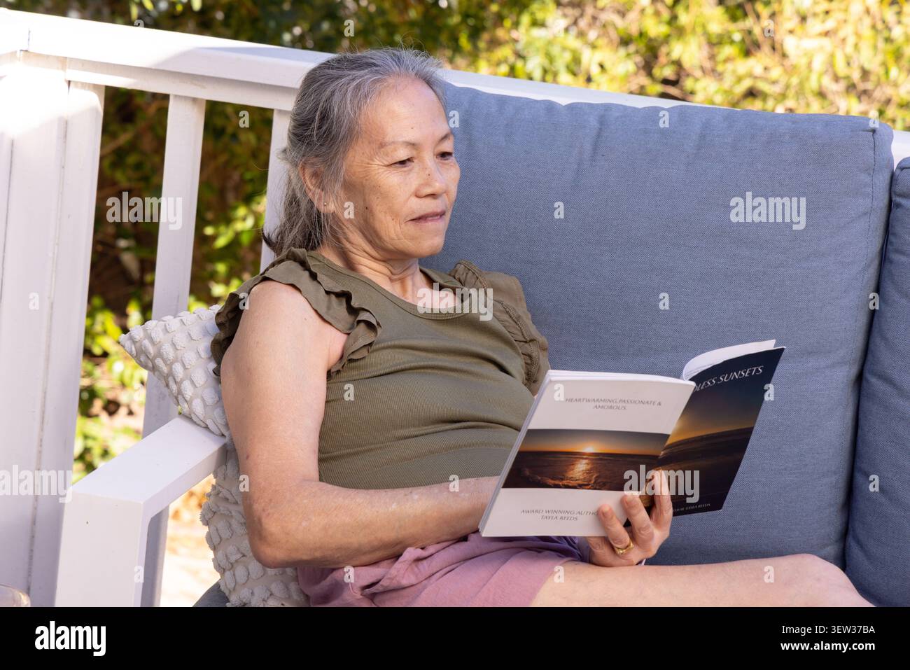 Senior Asian woman reclining on blue-gray porch sofa, leaning on pillow, reading paperback Stock Photo