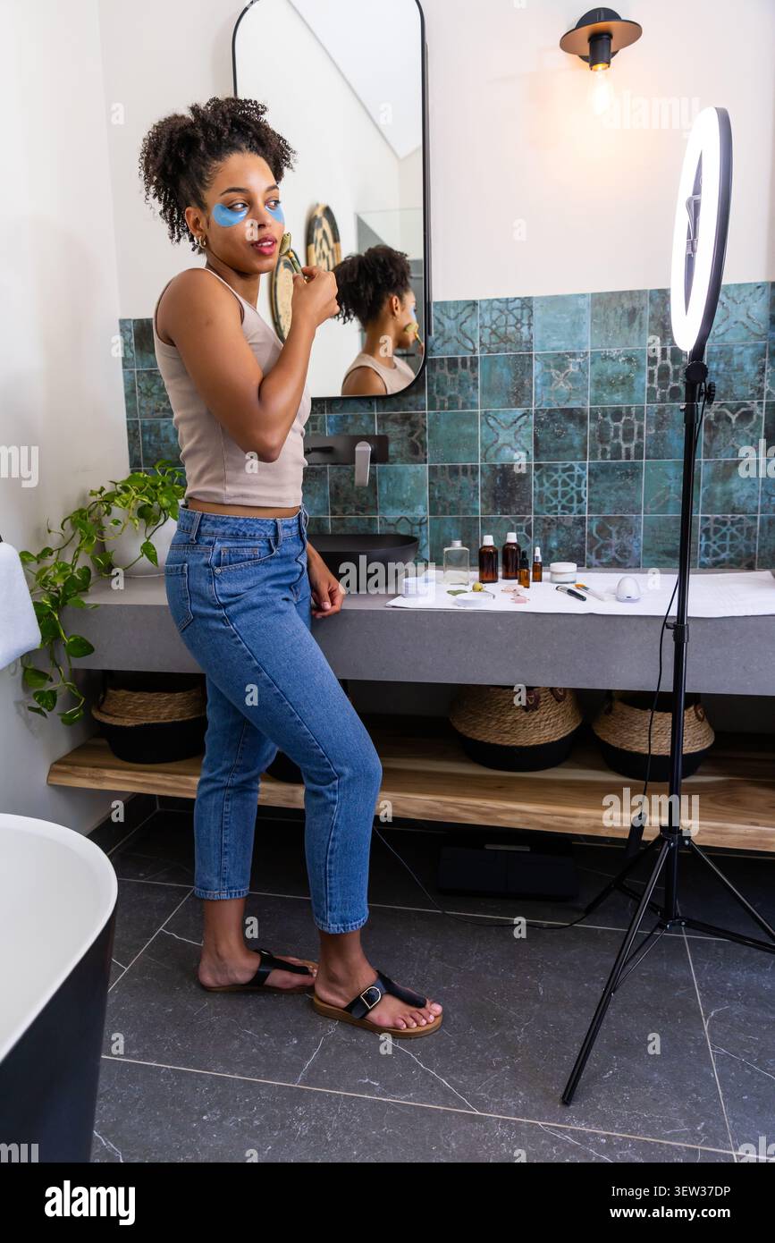 African American woman applying hydrogel eye patches with applicator at vanity by ring light Stock Photo