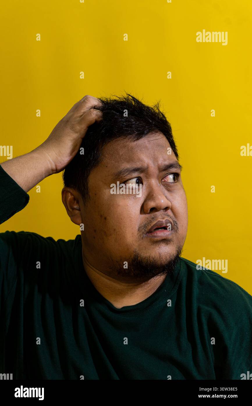 Confused bearded Asian male scratching his head while looking to the side with a skeptical expression against a solid bright yellow background. His fi Stock Photo