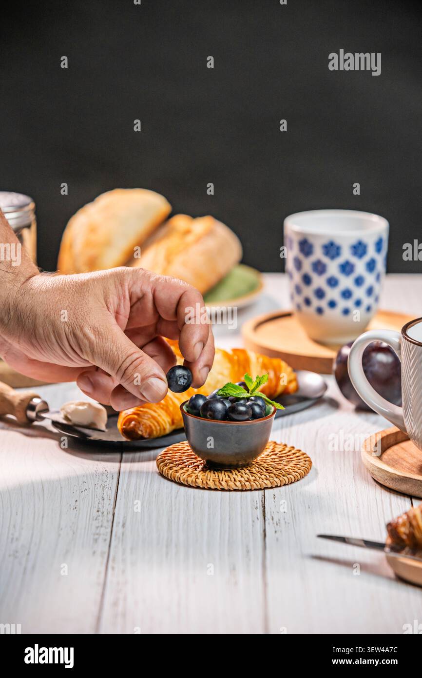 In the tranquil atmosphere of a morning, bathed in natural light, a hand carefully places blueberries, one by one, into a small, handcrafted ceramic b Stock Photo