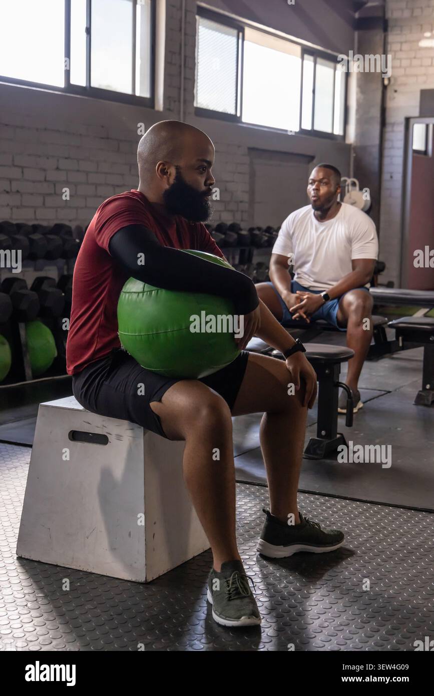 African men sitting on plyometric box holding green medicine ball at gym, wearing workout clothes Stock Photo