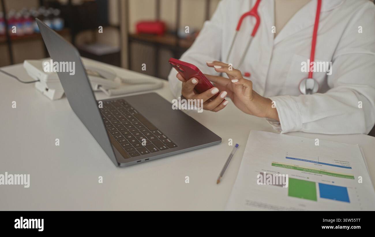 Woman doctor taps red smartphone with hands and stethoscope beside laptop on desk inside building; focus. Stock Photo