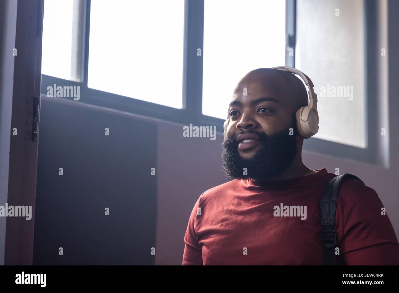 Man standing in corridor by windows wearing maroon tee, headphones and backpack, copy space Stock Photo