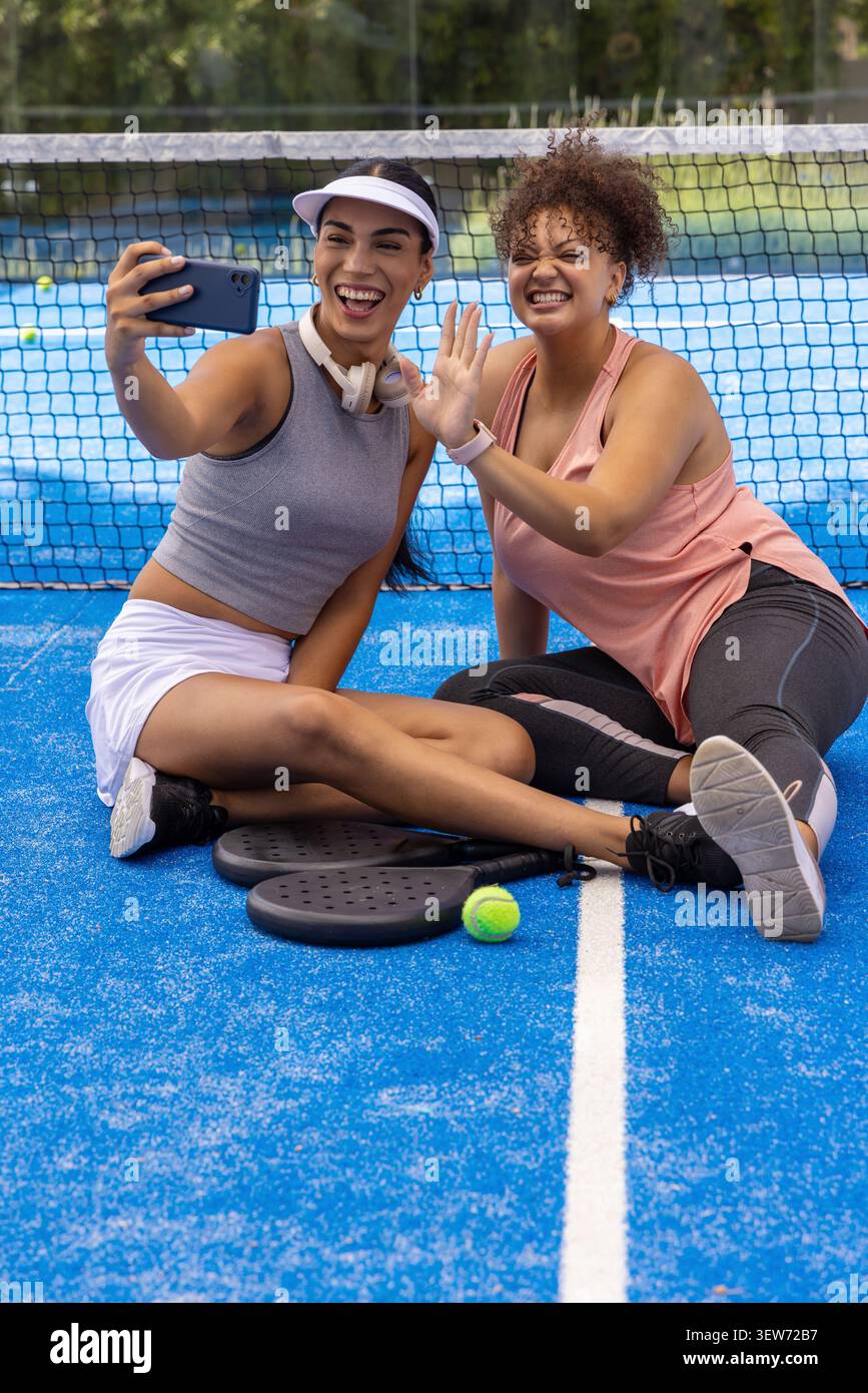 African American female duo taking smartphone selfie sitting on blue court with paddles and ball Stock Photo