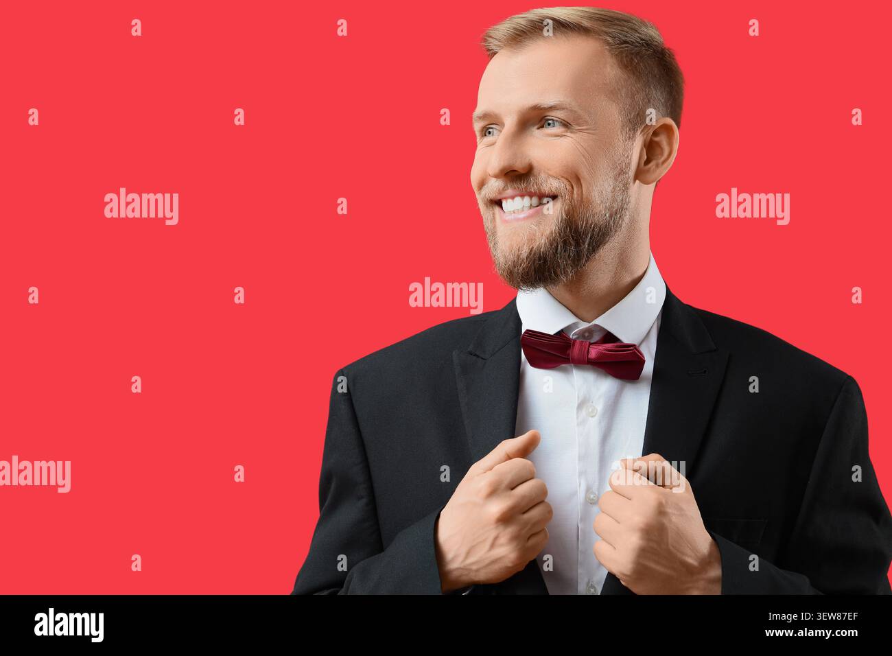Smiling man in tuxedo adjusting jacket on red background Stock Photo