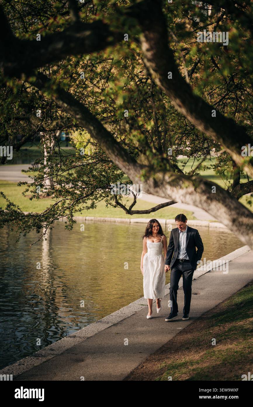 Elegant couple walks hand in hand by a park pond Stock Photo