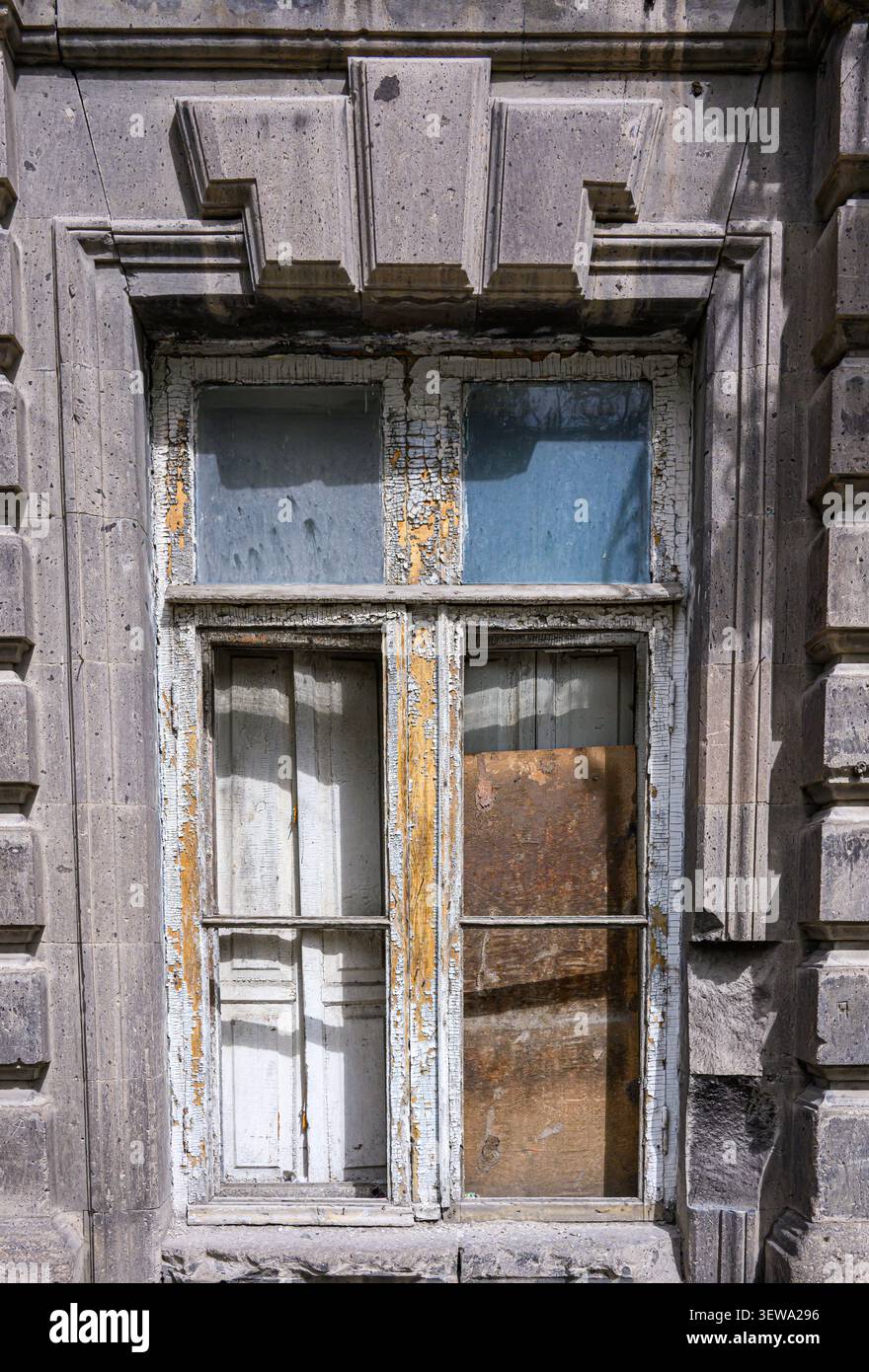 Old weathered window frame with peeling paint and broken glass. Stone wall surrounds worn wooden sash. Sunlight highlights decayed textures. Rust stai Stock Photo