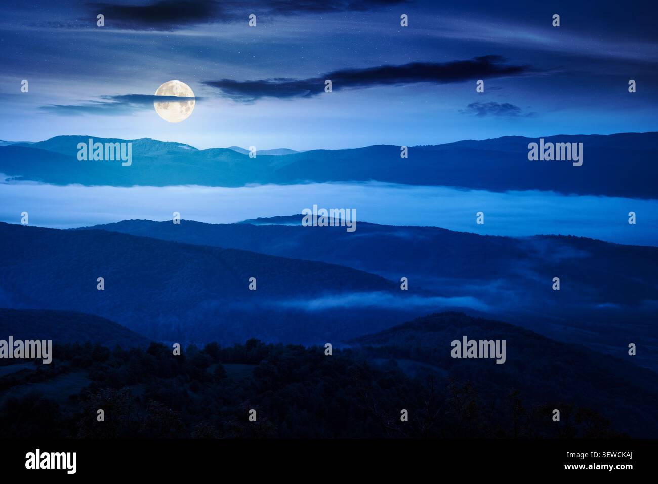 foggy carpathian countryside scenery in summer at night. mysterious view of mountainous landscape with rural fields on rolling hills. full moon over f Stock Photo