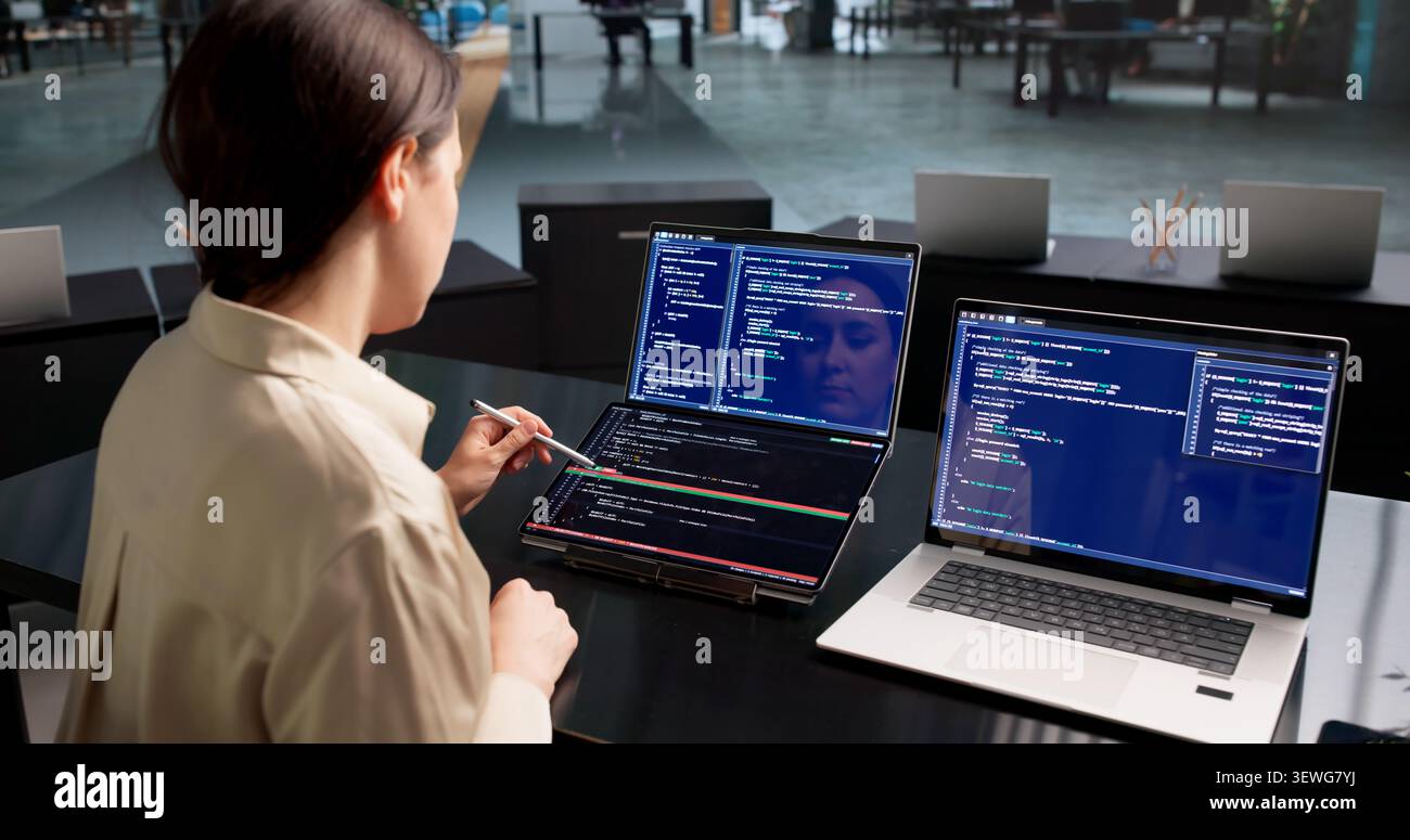 Young Female Software Developer Reviews Code Changes On Dual Touchscreen Laptops. Stock Photo