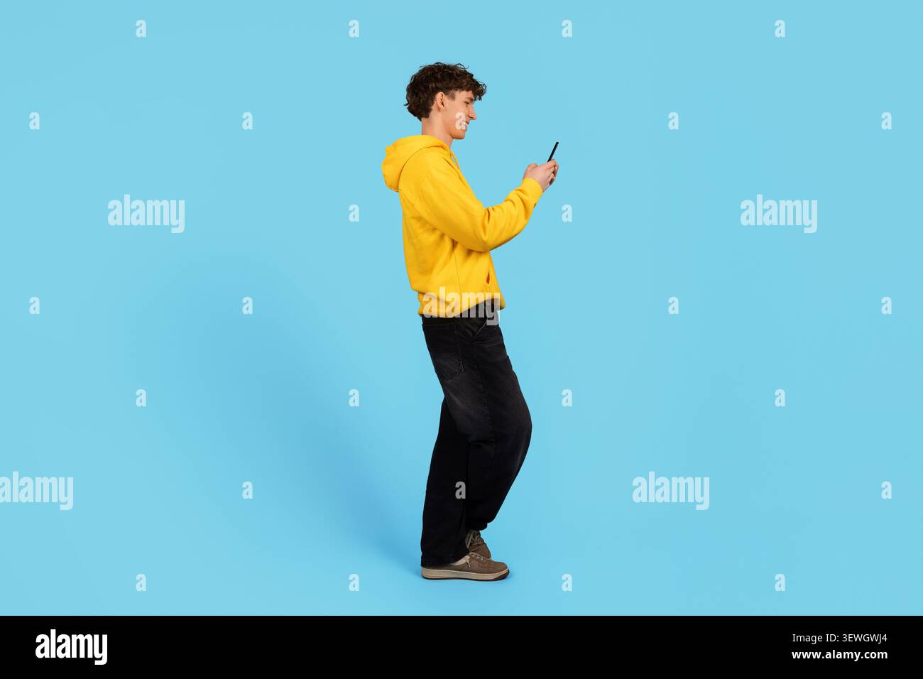 Young guy in yellow hoodie using smartphone on blue studio background Stock Photo