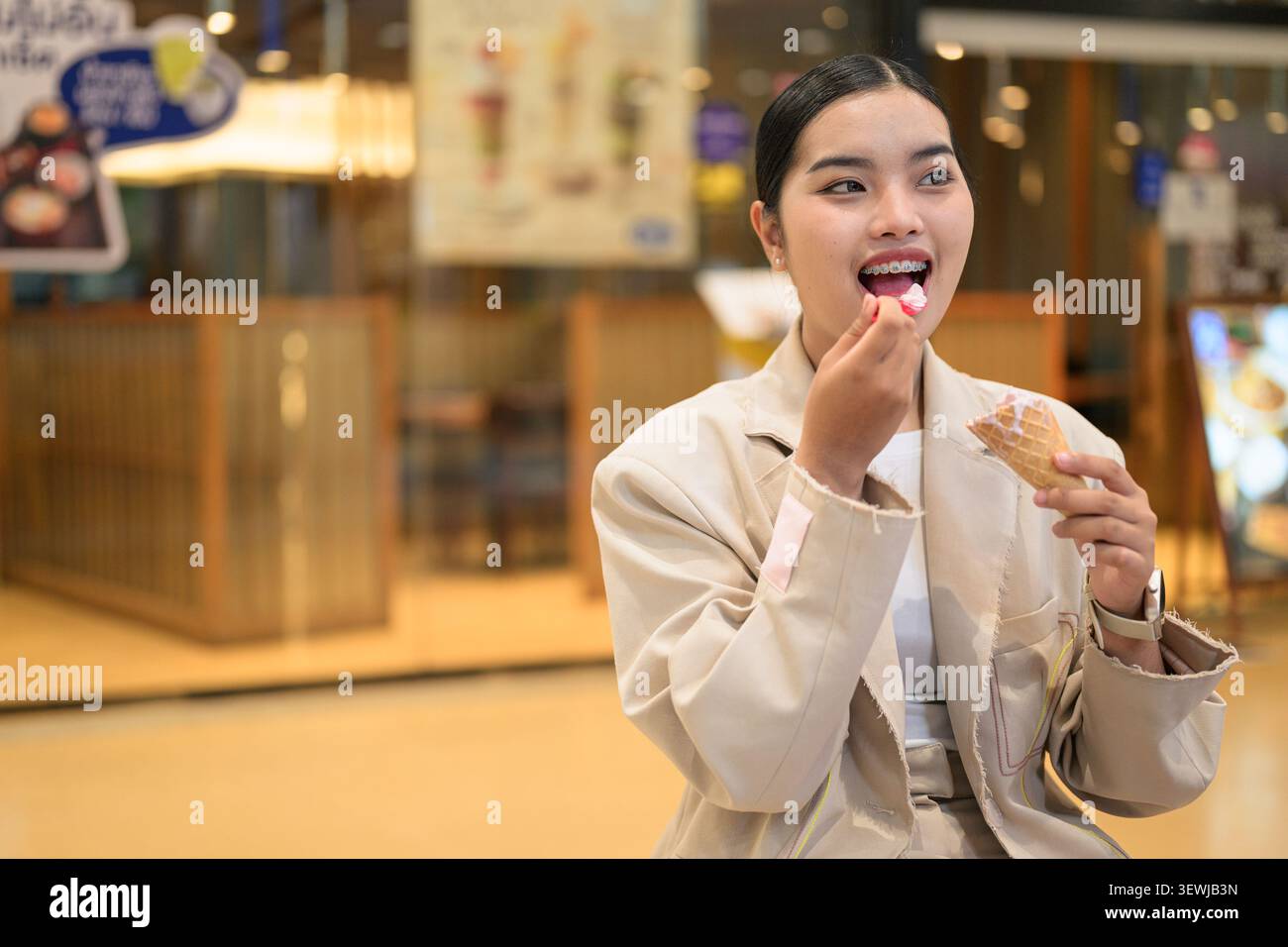 A cheerful young Southeast Asian Thai woman from the Isan region sitting inside a modern shopping mall while putting a small red spoon into her mouth Stock Photo
