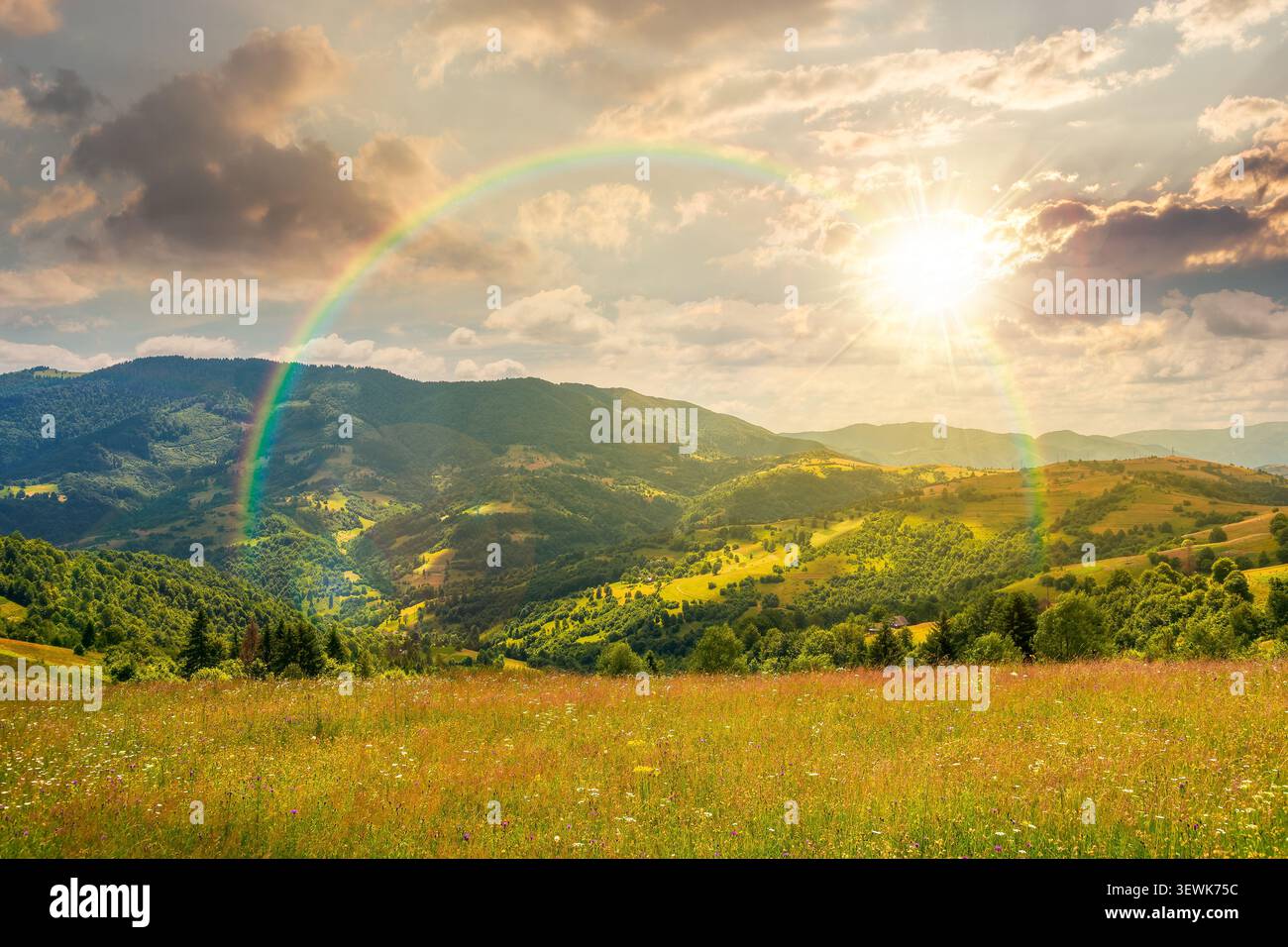 sunset over mountain landscape with grassy field. trees on the rolling hills and rural valley beneath a cloudy sky with rainbow in the background. vie Stock Photo