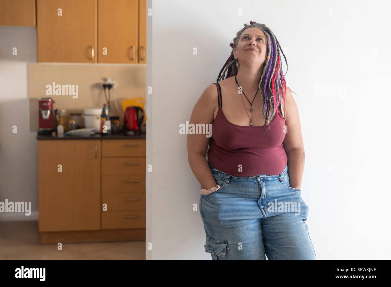 A middle-aged woman with colorful dreadlocks stands leaning against a white wall in her kitchen, looking up thoughtfully with a smile. Stock Photo