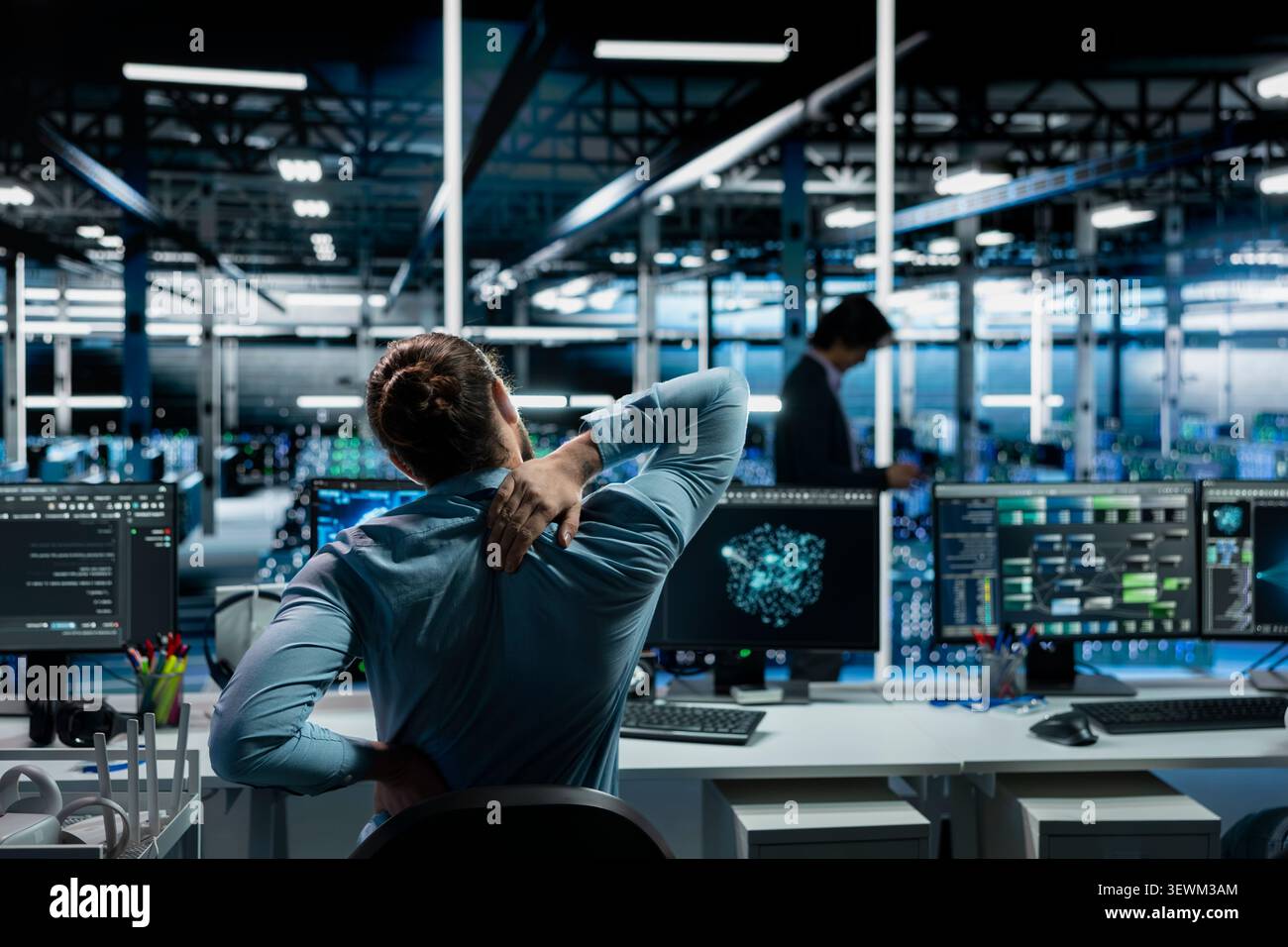 Software developer stretches at a workstation in a server room to relieve back fatigue after long technical work. Stock Photo
