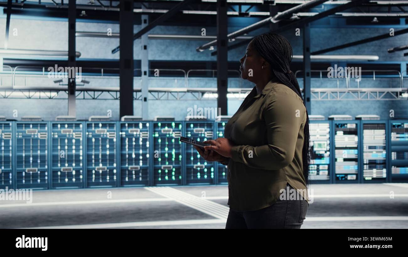 Data center technician monitors operational metrics on tablet inside a server room to improve infrastructure performance. Stock Photo