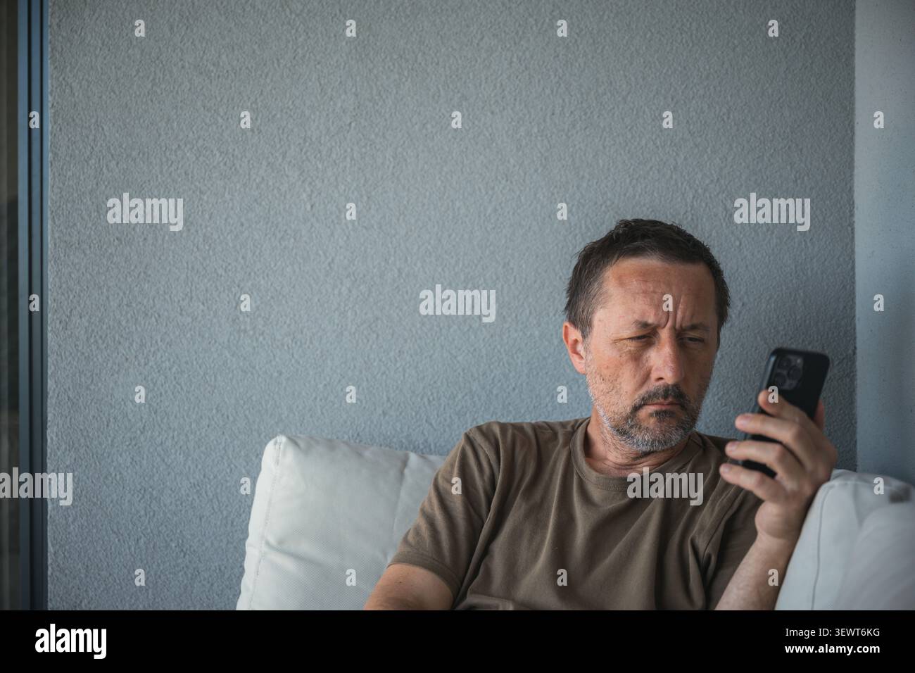 Middle-aged man looking thoughtfully at smartphone while sitting on sofa, selective focus. Stock Photo