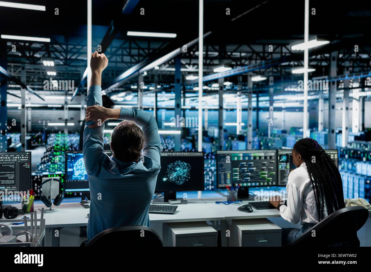 Software developer stretches at a workstation in a server hub to relieve back fatigue after long technical work. Stock Photo