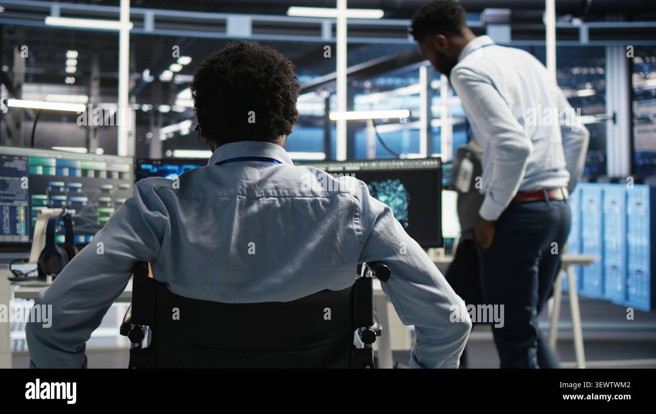 Data infrastructure center software programmer in wheelchair arriving at the office operates AI visual programming workflow to minimize system downtime. Stock Photo