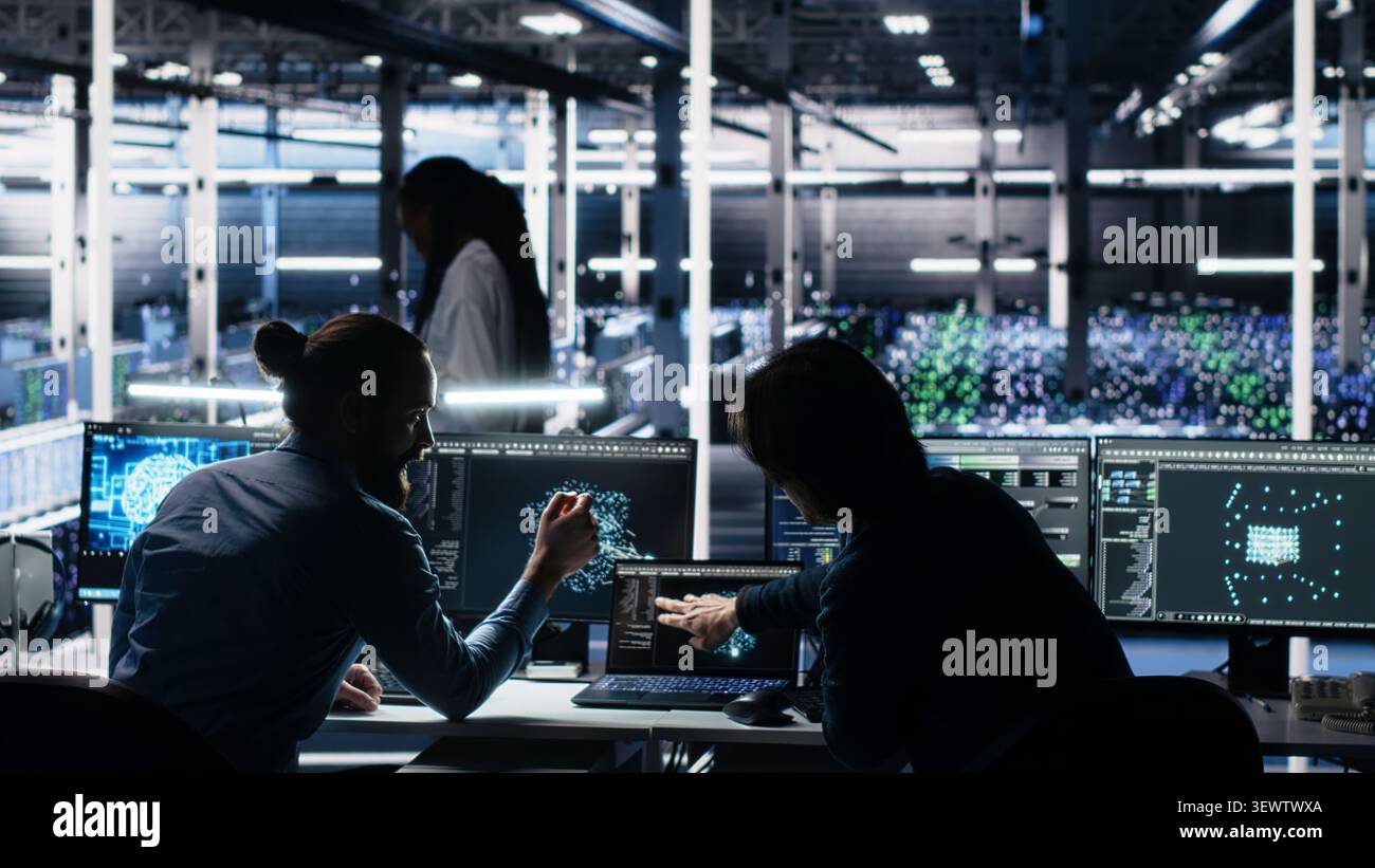 Senior programmer and coworker programs on a computer workstation in a data center with generative AI assistance. Stock Photo