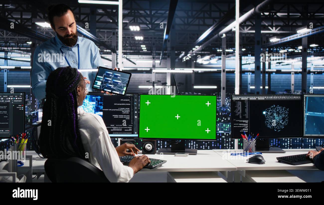 Female software programmer talking with team supervisor in high tech server room, checks AI diagnostics on chroma key computer. Stock Photo