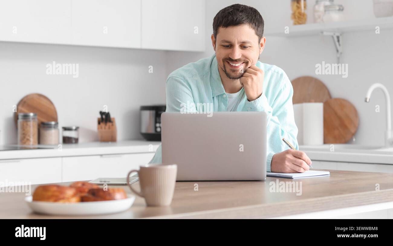 Man working on laptop and taking notes in kitchen Stock Photo