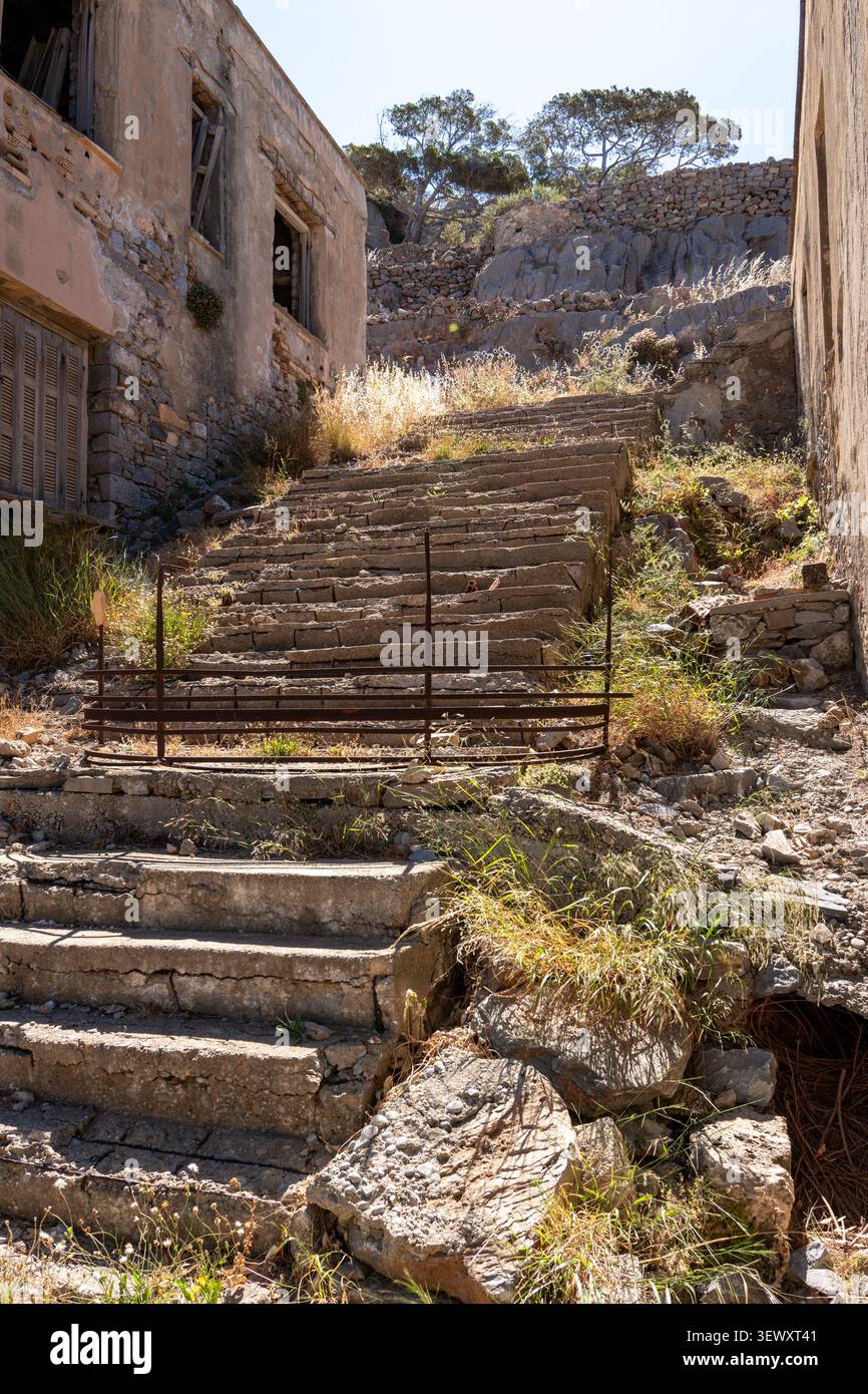 Weathered concrete staircase with metal railing stands between ruined stone buildings with green trees on hill under clear sky on Spinalonga island Cr Stock Photo