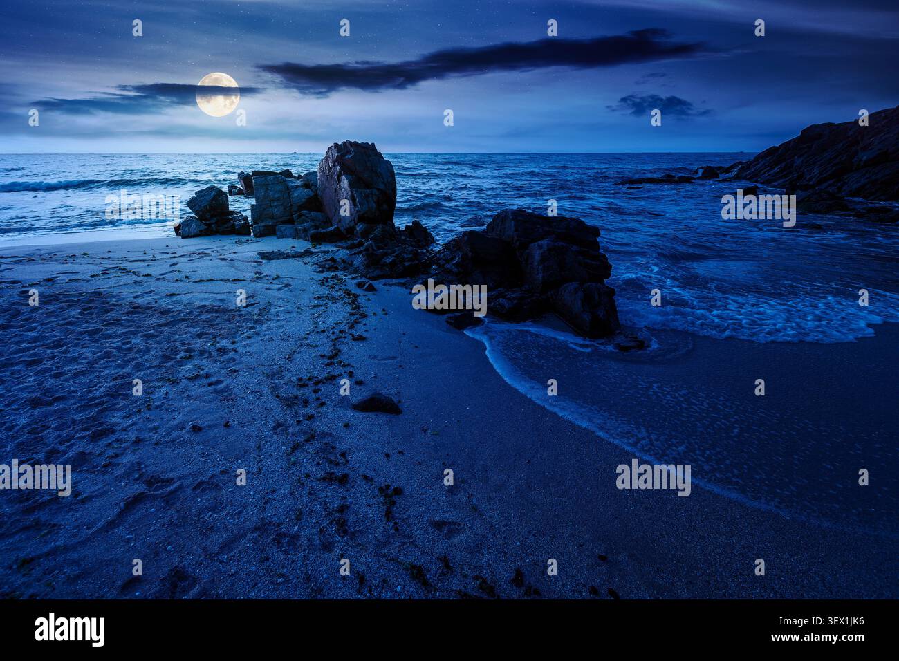 huge stones on the sandy beach at night. mysterious holiday season background at the sea. full moon over clouds on the dark sky above horizon. view of Stock Photo