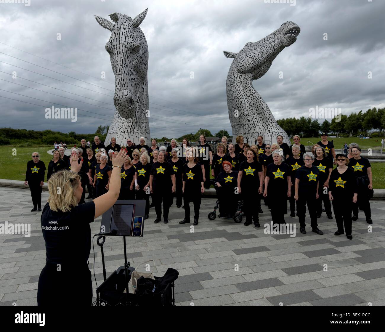 Rock Choir members perform beneath The Kelpies at Helix Park, Falkirk, conducted by a blonde female choir leader on 27 June 2026 Stock Photo