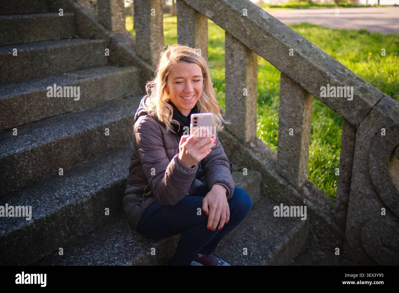 Woman sitting on old stone stairs in a park, smiling while looking at her smartphone Stock Photo