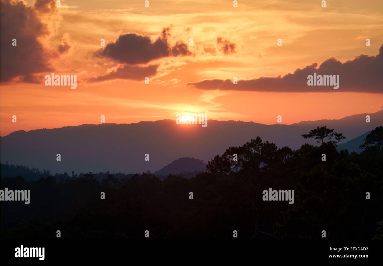 Vibrant orange sunset with clouds over layered mountain ridge with silhouetted trees in tropical forest Stock Photo