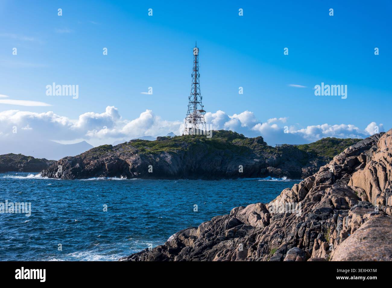 Tall red and white communication tower stands on a rocky, vegetation-covered island in Lofoten. Dramatic coastal landscape with deep blue Stock Photo