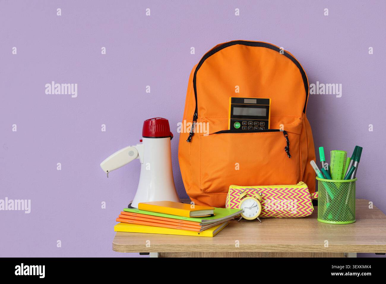 Orange backpack with school supplies on wooden table against purple wall Stock Photo