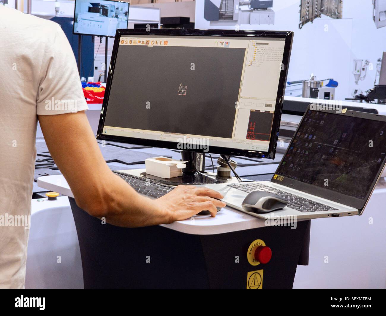 An operator uses specialized CAD/CAM software on a dual-screen computer setup to control an industrial CNC cutting machine in a modern manufacturing f Stock Photo