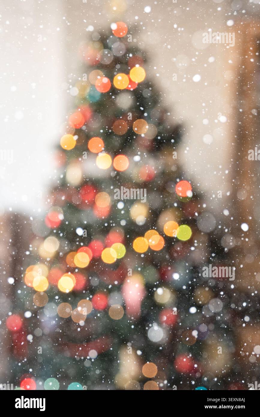Vertical festive background featuring colorful Christmas tree bokeh lights in a warm home interior. A soft overlay of falling snowflakes adds a magica Stock Photo