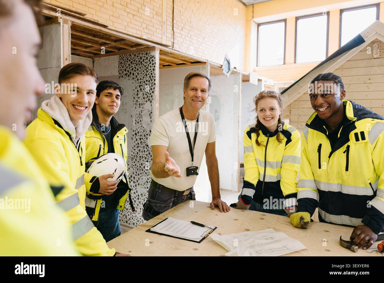Smiling instructor gesturing while standing with vocational students around workbench inside construction workshop at up Stock Photo