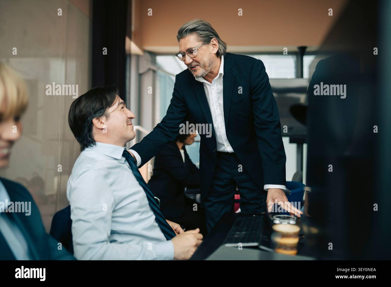 Senior businessman talking with hand on shoulder of male colleague sitting at desk in office Stock Photo