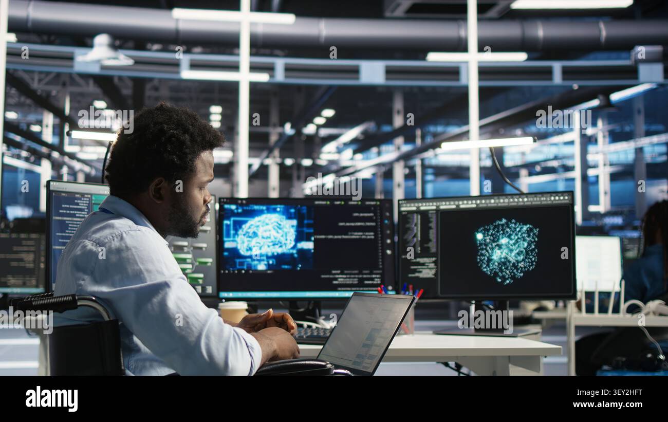 Data infrastructure center computer scientist in wheelchair working with AI visual programming system to minimize system downtime, while with disability working with AI visual programming tool. Stock Photo