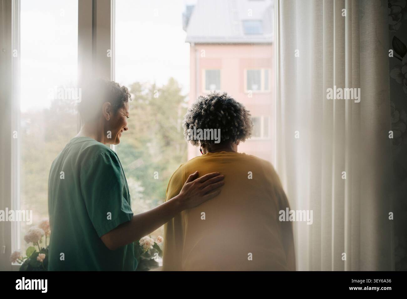 Smiling female nurse next to senior woman looking out of window Stock Photo