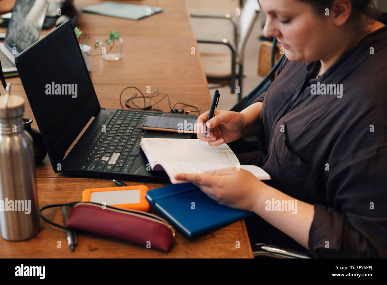Female freelancer taking down notes in diary while sitting with laptop at shared working desk Stock Photo